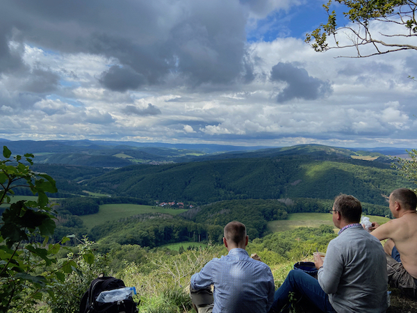 Drei Wanderer pausieren sitzend, auf einer Anhöhe. Sie blicken auf eine bewaldete Hügellandschaft und blau/bewölkten Himmel.